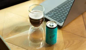 A cold brew coffee can next to a laptop and a glass with cold brew coffee.